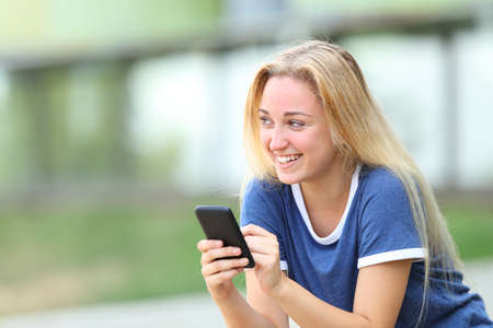 Happy teenage girl holding phone looking at sidesitting in an university campusの写真素材