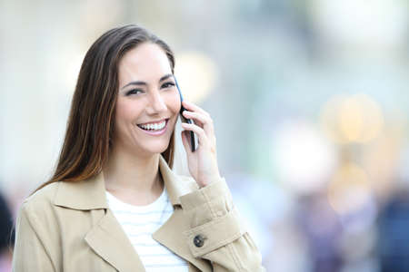 Happy woman talks on smart phone and looks at camera standing in the street
