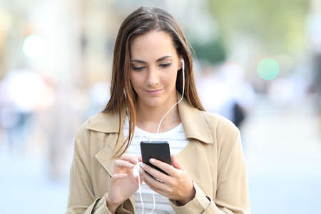Front view of serious girl wearing earphones listening to music on smart phone walking towards camera in the streetの写真素材