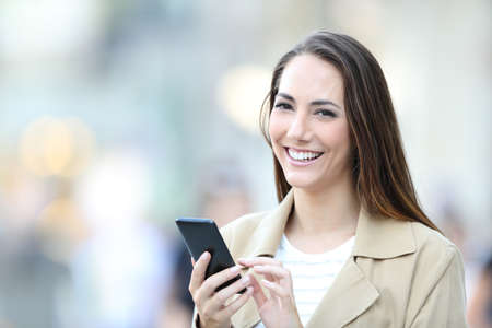 Happy woman holds a smart phone looking at camera standing in the streetの写真素材