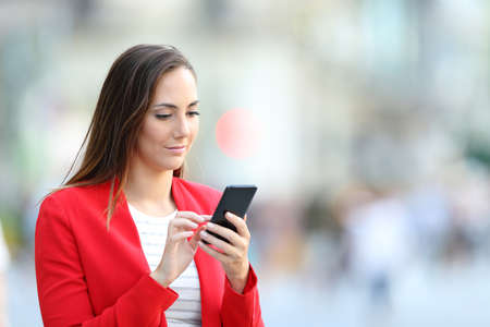 Serious elegant woman in red using smart phone standing in the streetの写真素材