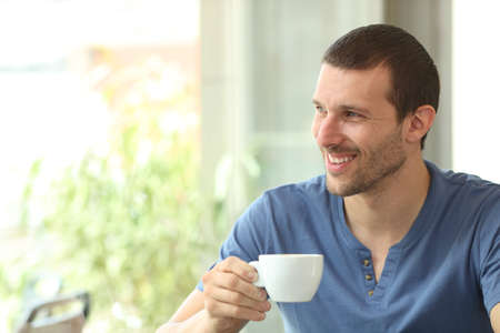 Happy man holds a coffee cup looking at side through a window in a coffee shop or homeの写真素材