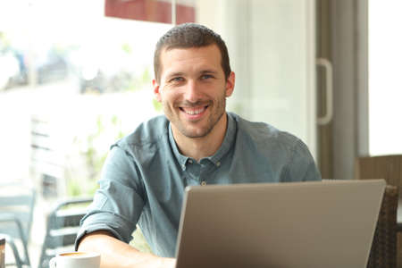 Happy man with a laptop posing looking at camera sitting in a coffee shopの写真素材