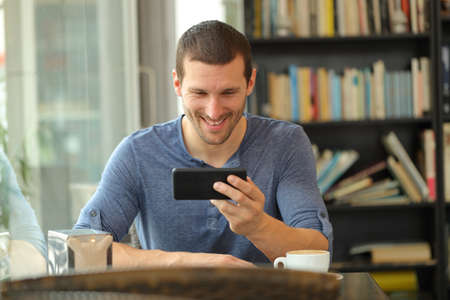 Front view portrait of a happy man watching media on horizontal smart phone sitting in a barの写真素材