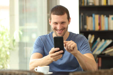 Front view of a happy man using smart phone sitting in a coffee shop or homeの写真素材