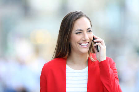 Front view portrait of a happy woman in red talking on phone walking towards camera in the streetの写真素材
