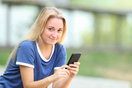 Relaxed teenage girl holding phone looking at you sitting in an university campusの写真素材