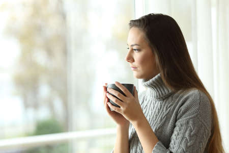 Worried woman holding a coffee mug looks through a window at home in winterの写真素材