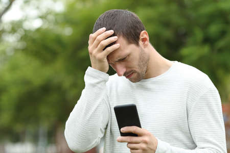 Sad adult man checking smart phone message complaining alone in a parkの写真素材