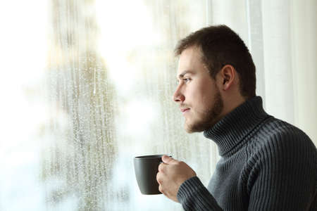 Side view portrait of a serious man holding a coffee mug looking the rain through a window at homeの写真素材