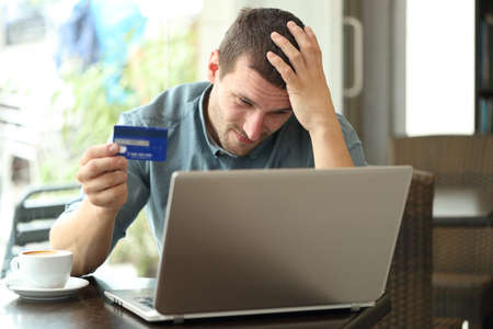 Worried man having problems paying with credit card and laptop sitting in a coffee shopの写真素材