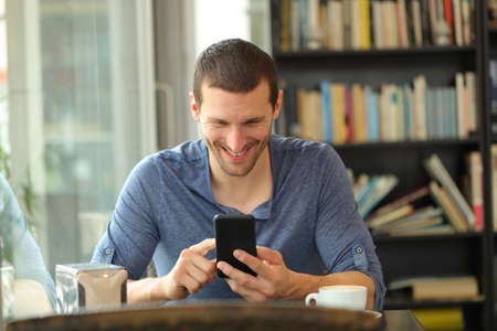 Front view portrait of a happy man using smart phone sitting in a coffee shop or homeの写真素材