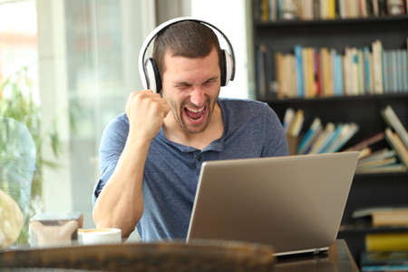 Excited man wearing headphones checking laptop celebrating good news sitting in a coffee shopの写真素材