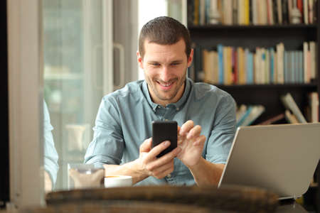 Front view portrait of a happy man using smart phone and laptop sitting in a coffee shop or homeの写真素材