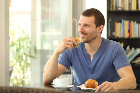 Happy man eating a croissant for breakfast sitting in a coffee shop looking through a windowの写真素材