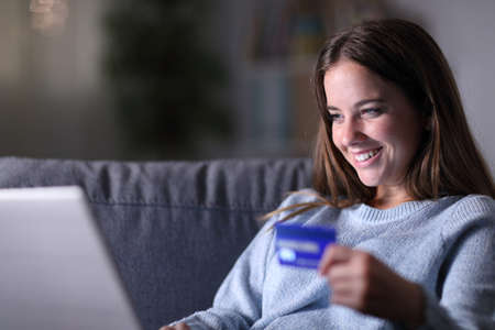 Happy woman using laptop and credit card to buy online sitting on a couch in the living room in the night at homeの写真素材