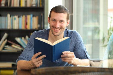 Front view portrait of a happy man holding a paper book posing looking at camera sitting in a coffee shop or homeの写真素材