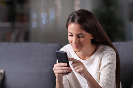 Serious girl uses a mobile phone sitting on a couch in the night at homeの写真素材