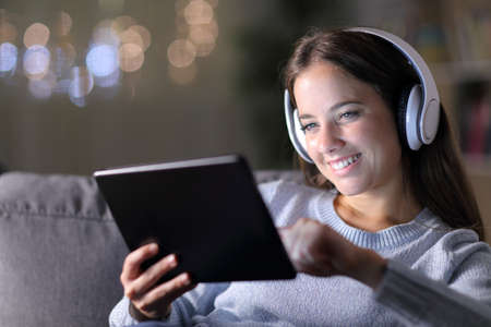 Happy woman wearing headphones uses a tablet sitting on a couch in the night at homeの写真素材