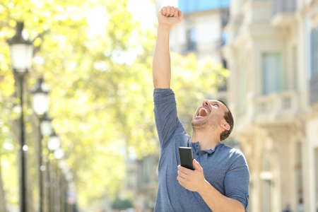 Excited adult man holding mobile phone celebrating success raising arm in the streetの写真素材