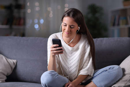 Relaxed girl listening to music on phone sitting on a couch in the night at homeの写真素材