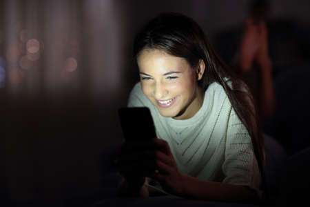 Happy girl checking phone in the night with the face lit lying on a couch in the night at homeの写真素材