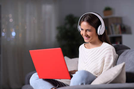 Happy girl wearing headphones using a red laptop sitting on a couch in the night at homeの写真素材