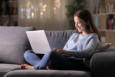Full body portrait of a happy woman using a laptop sitting on a couch in the night at homeの写真素材