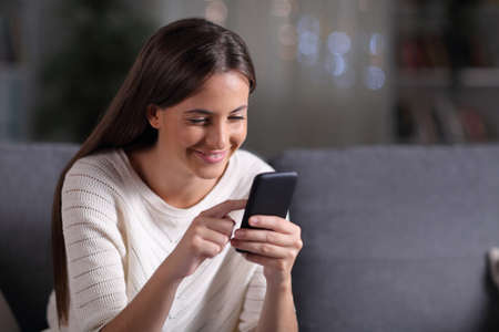 Happy teen browsing mobile phone content sitting on a couch in the night at homeの写真素材