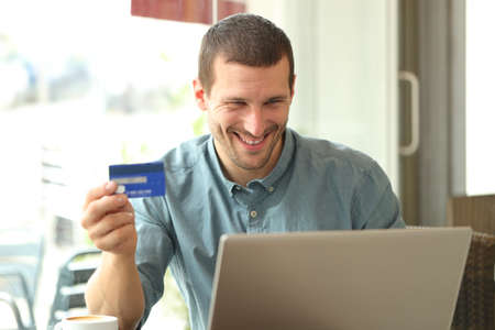 Happy adult man buying online with credit card and laptop sitting in a coffee shopの写真素材