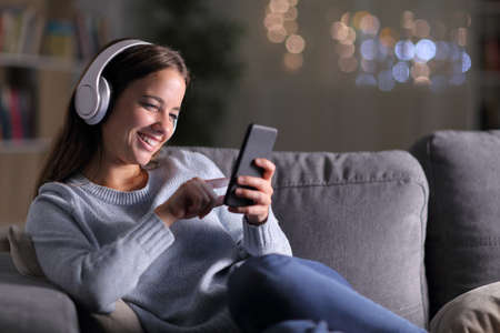 Happy woman listening to music using smart phone sitting on a couch in the night at homeの写真素材