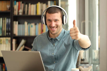 Happy adult man with laptop and headphones gesturing thumbs up sitting in a coffee shopの写真素材