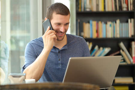 Happy man talks on phone checking laptop content sitting in a bar or homeの写真素材
