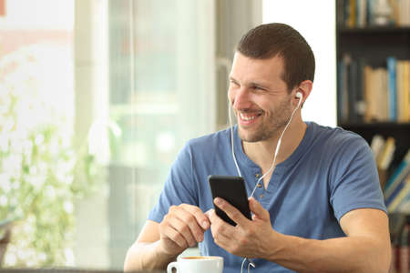 Happy man listening to music using smart phone looking outdoors through a window sitting in a bar or homeの写真素材