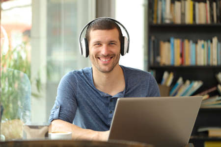 Happy man posing with headphones and laptop looking at camera sitting in a coffee shopの写真素材