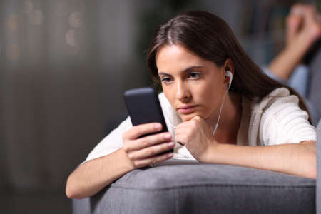 Serious girl listening to music reading mobile phone content lying on a couch in the night at homeの写真素材