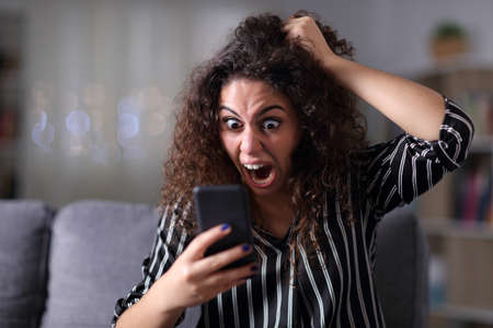 Hysterical woman yelling checking smart phone content sitting on a couch in the night at homeの写真素材