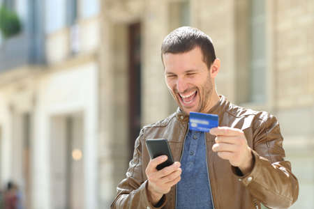Excited man paying on line with credit card and mobile phone in the streetの写真素材