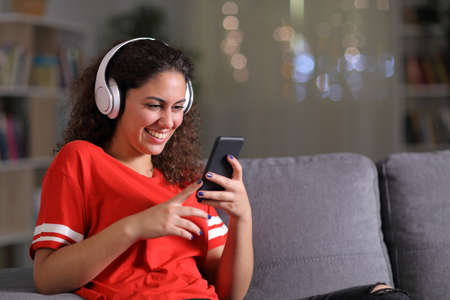 Happy girl in red listening to music checking smart phone sitting on a couch in the living room in the night at homeの写真素材