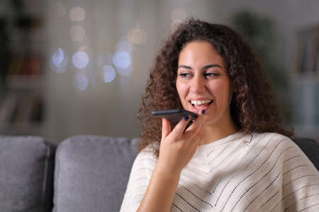 Happy woman using voice recognition on mobile phone sitting on a couch in the living room in the night at homeの写真素材