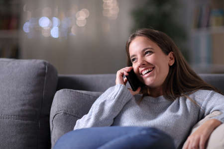 Happy woman talking on mobile phone sitting on a couch in the living room in the night at homeの写真素材