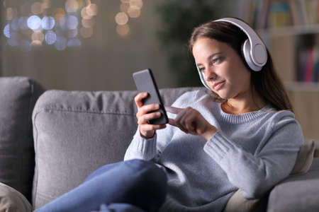 Woman listening to music using headphones and mobile phone sitting on a couch in the living room in the night at homeの写真素材
