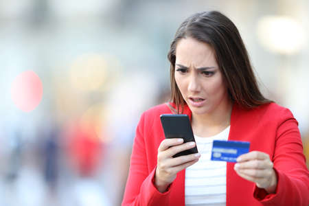 Worried woman holding credit card looking at smart phone in the streetの写真素材