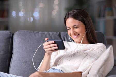 Happy girl watching media on smart phone sitting on a couch in the night at homeの写真素材