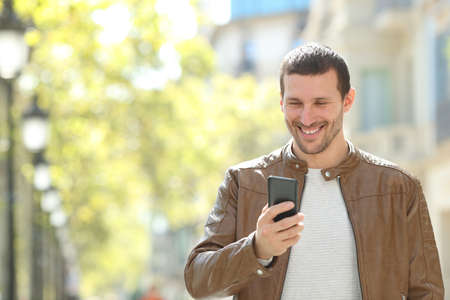 Front view portrait of a happy adult man using a smart phone walking in the streetの写真素材