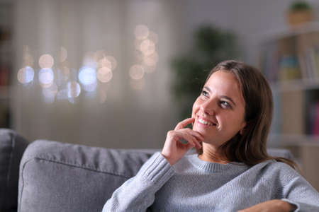 Happy woman thinking looking at side sitting on a couch in the living room in the night at homeの写真素材