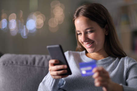 Happy woman buying with mobile phone and credit card sitting on a couch in the living room in the night at homeの写真素材