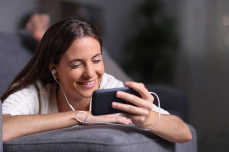 Happy girl watching movie on smart phone lying on a couch in the night at homeの写真素材