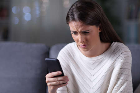 Worried girl checking smart phone news sitting on a couch in the night at homeの写真素材