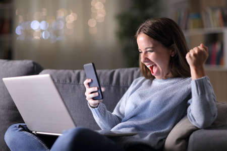 Excited woman using phone and laptop sitting on a couch in the living room in the night at homeの写真素材